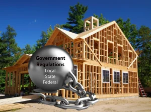 Framed wooden house under construction with a heavy ball and chain lableled "Government Regulations - Local, State, Federal symbolizing the regulatory burdens on housing costs